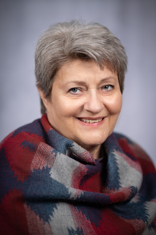 Porträtaufnahme einer lächelnden Frau mit kurzen, grauen Haaren, die einen gemusterten Schal in Rot-, Blau- und Grautönen trägt.